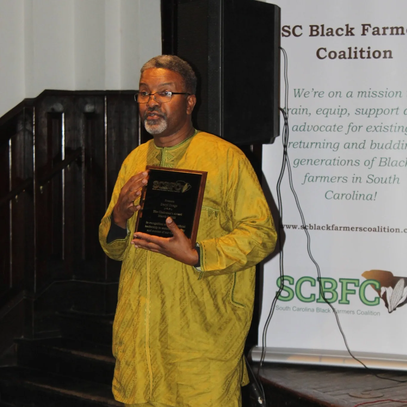 A speaker holds an award beside a South Carolina Black Farmers Coalition banner.