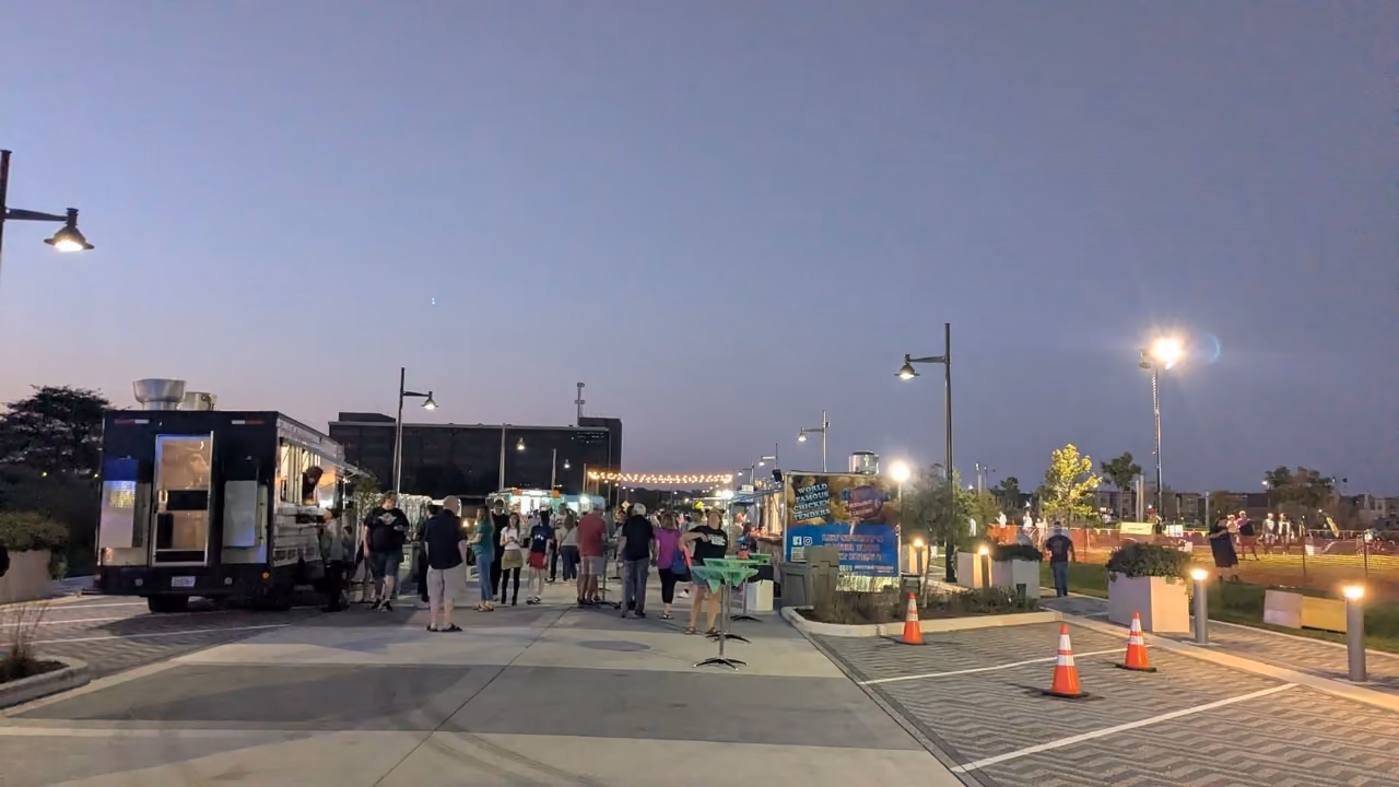 Food trucks and a crowd along Promenade Drive at dusk, string lights and lamp posts overhead