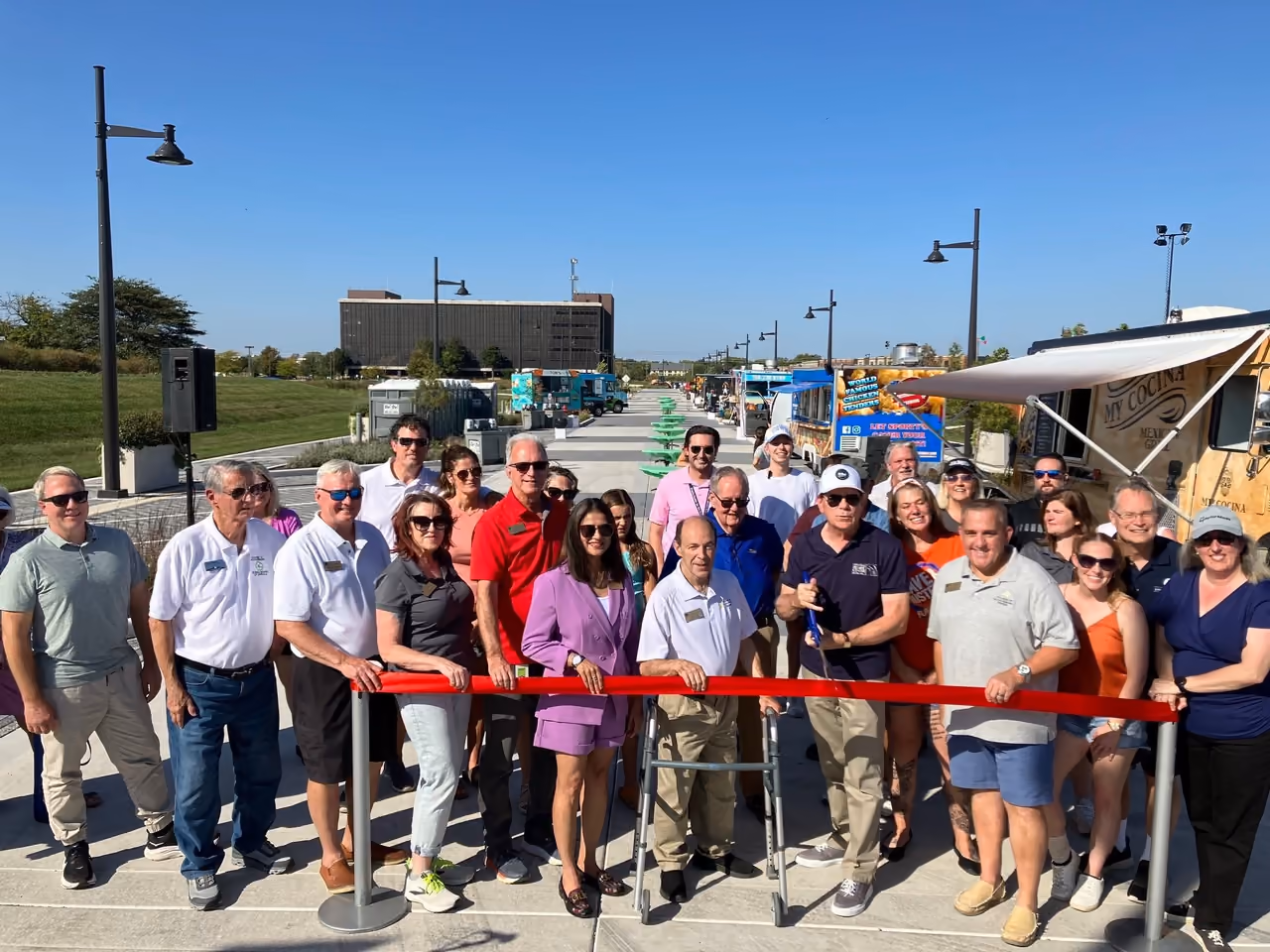 A crowd holds a long red ribbon across Promenade Drive at the opening of 90 North Park, food trucks lined up behind them