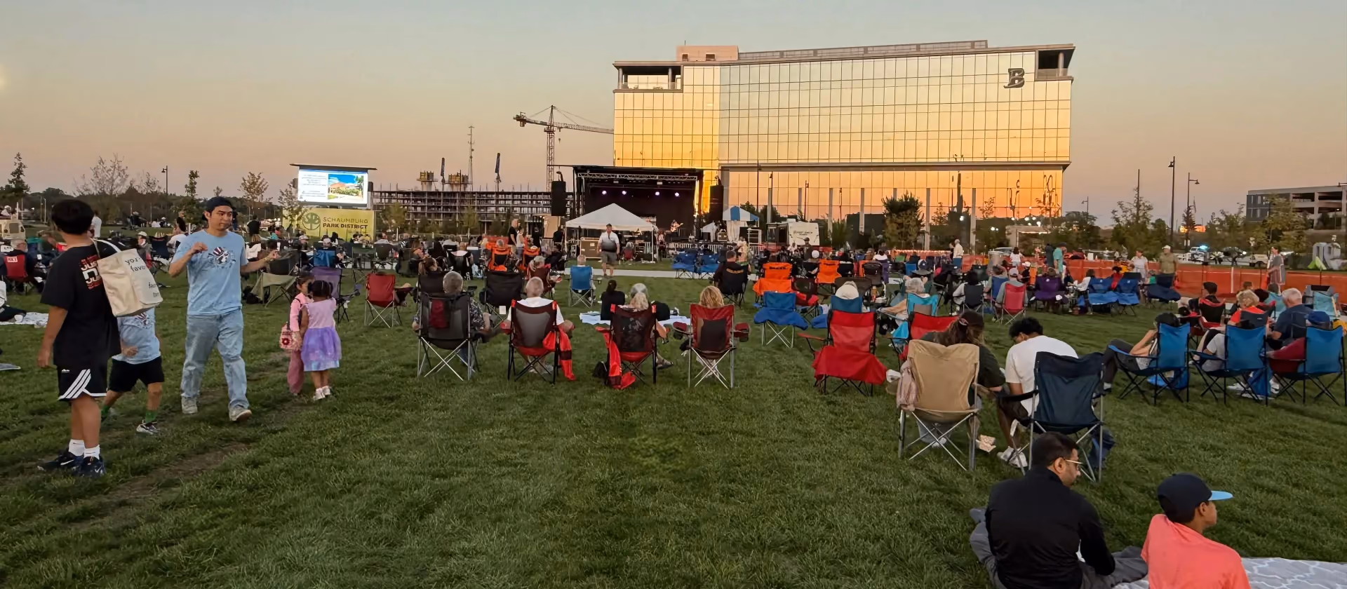 Families on lawn chairs and blankets at Schaumburg Live! @ 90N, the sun setting on the glass Boler Company building behind the stage