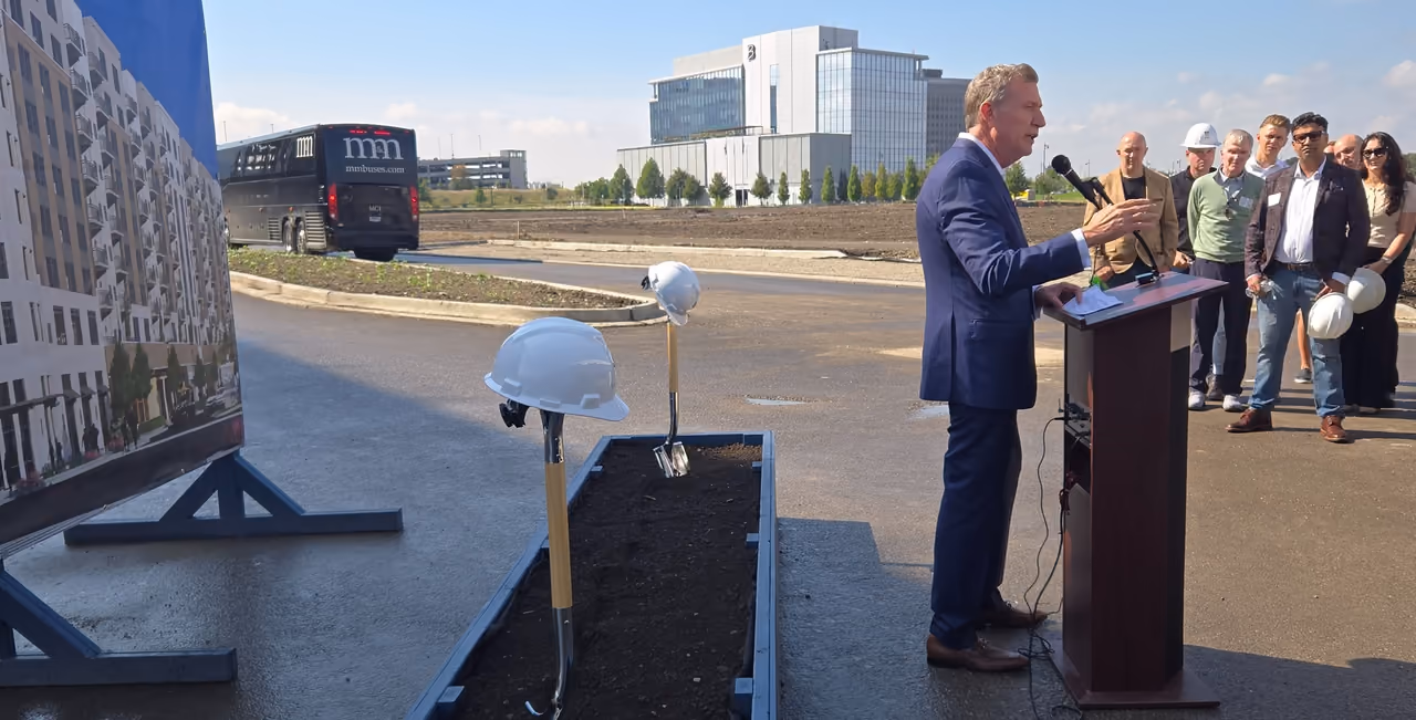 A speaker at a podium at The District at Veridian groundbreaking, with hard hats and ceremonial shovels set out in front