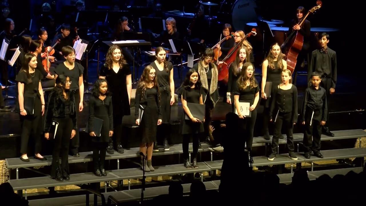 Young singers in black on choir risers on the Maggie Atcher Theatre stage, the youth orchestra behind them