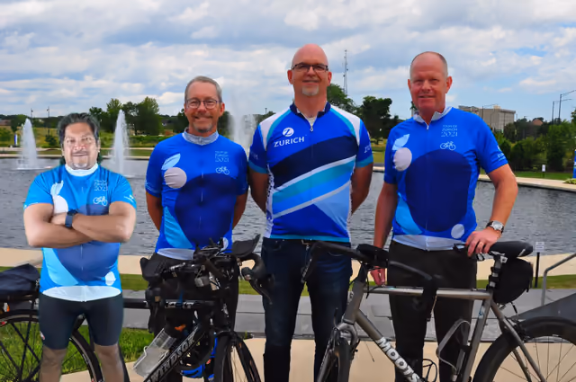 Four cyclists in team jerseys standing with their bikes beside a pond with fountains