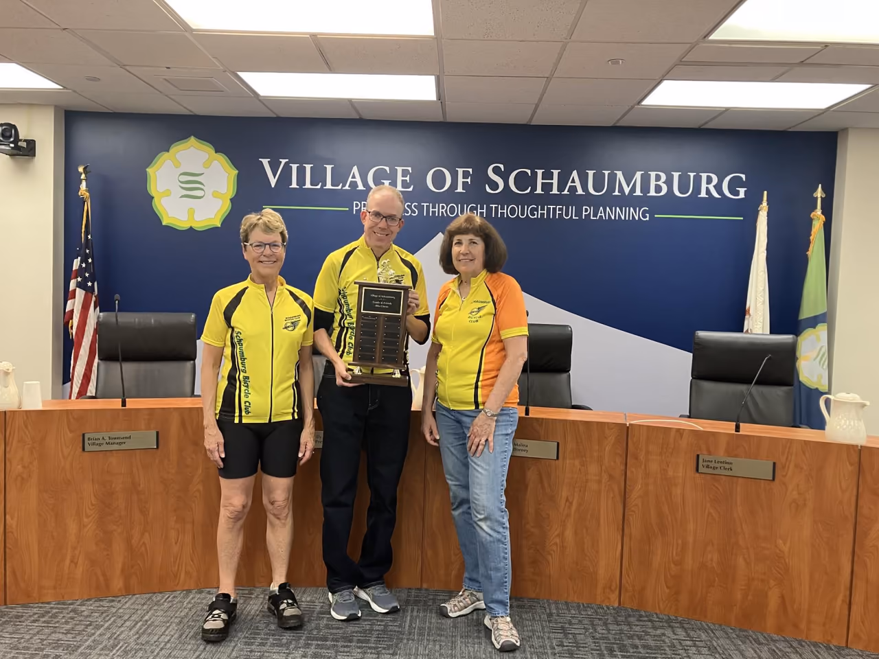 Three Bike Month riders holding an award in the Village Board room, the Village seal and motto on the wall behind them