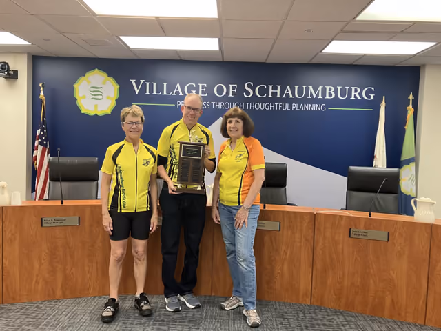Three Bike Month riders holding an award in the Village Board room, the Village seal and motto on the wall behind them