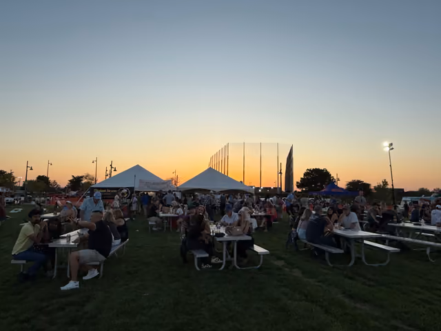 People at picnic tables on the 90 North Park lawn at sunset, white event tents behind them