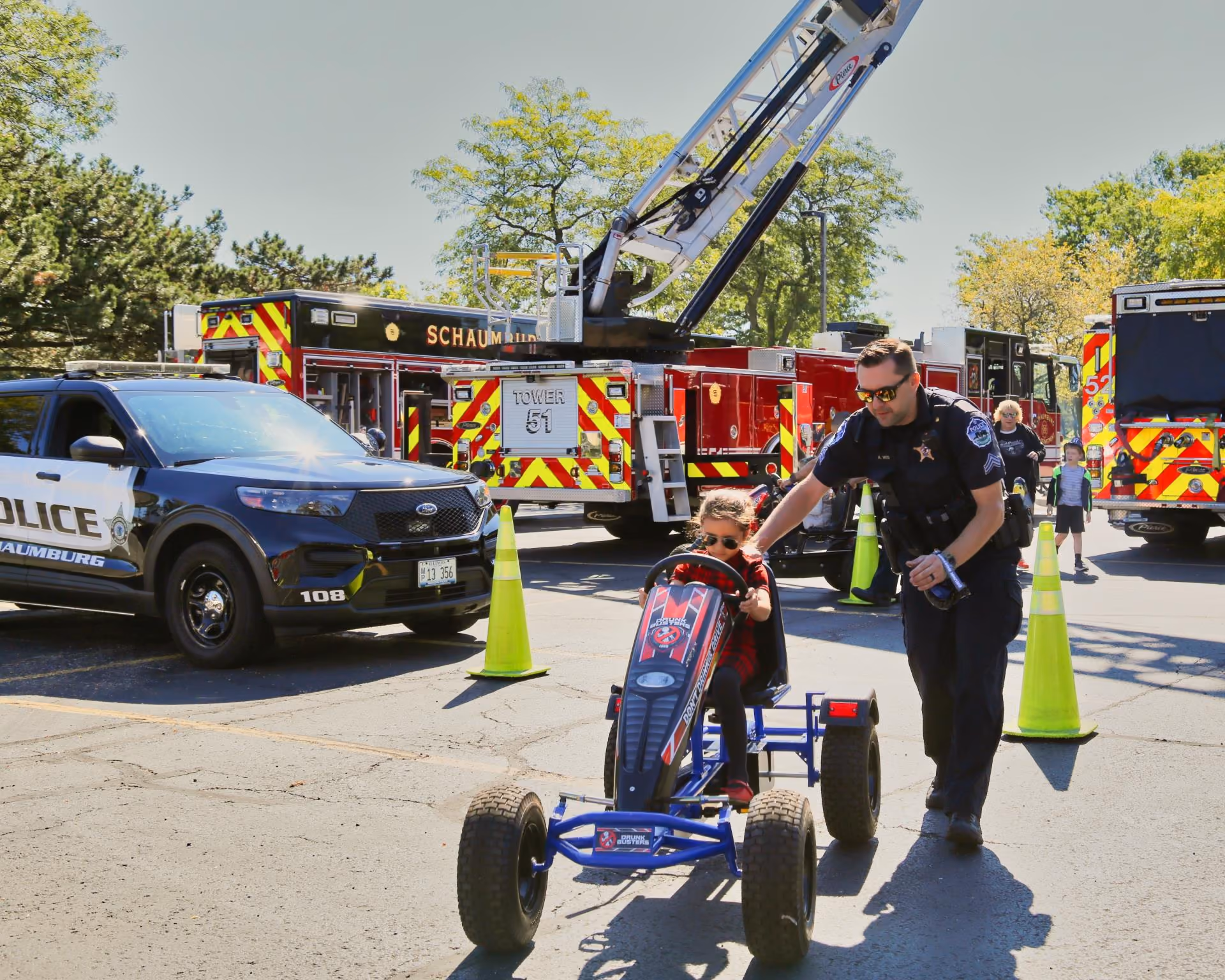 A police officer walks beside a child riding a pedal tractor past fire engines and a squad car at the Public Safety Open House
