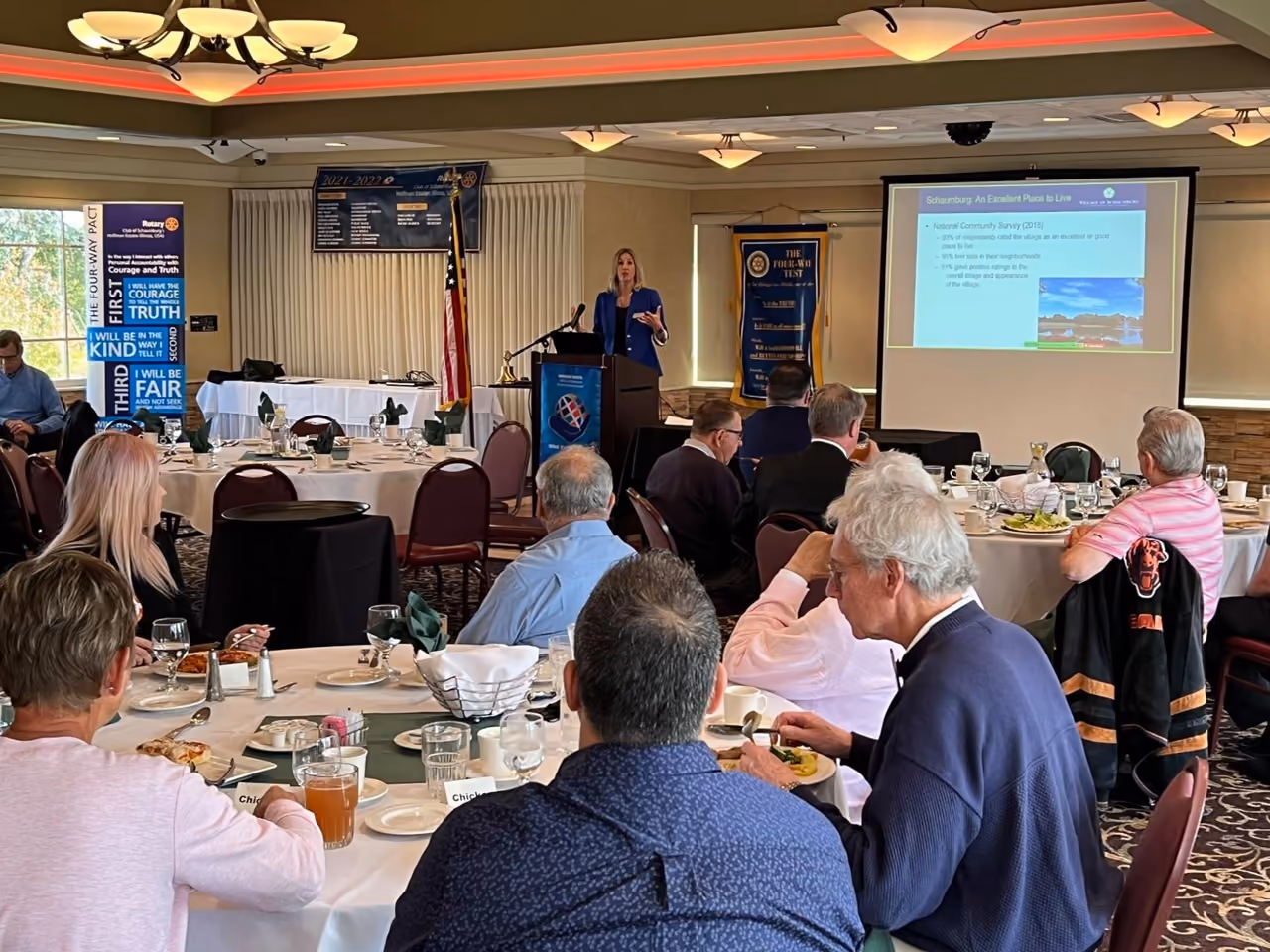 A presenter at a lectern beside a projection screen, speaking to a Rotary Club seated at round tables