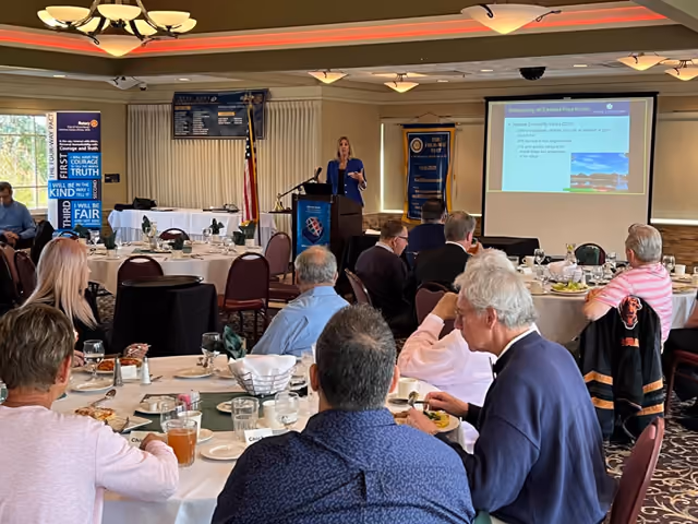 A presenter at a lectern beside a projection screen, speaking to a Rotary Club seated at round tables