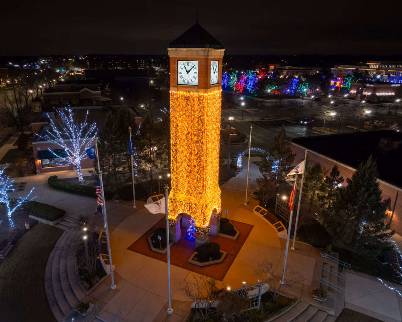 A clock tower wrapped in golden lights above a plaza at night during the tree lighting
