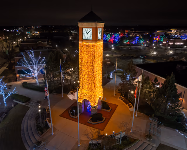 A clock tower wrapped in golden lights above a plaza at night during the tree lighting