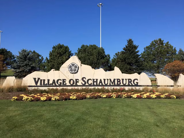 A Village of Schaumburg welcome sign with the emblem, flower beds and evergreens behind it