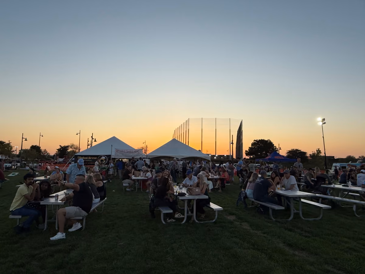 People at picnic tables on the 90 North Park lawn at dusk, event tents behind them