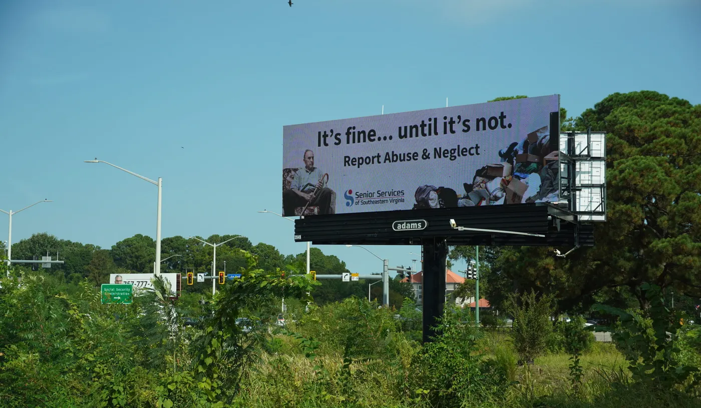A Senior Services elder-abuse awareness billboard beside a wooded Hampton Roads highway, urging drivers to report abuse and neglect.