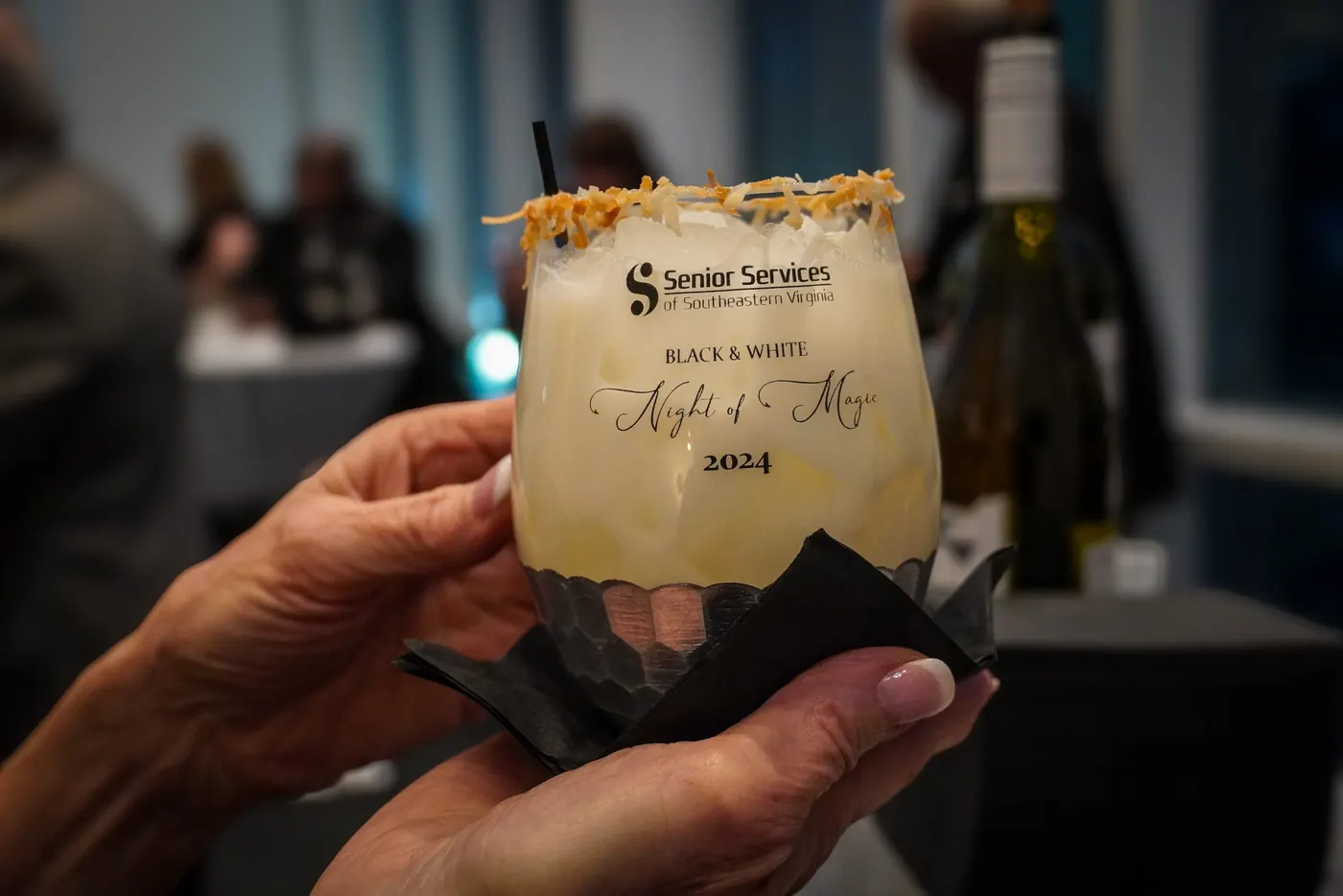 Hands holding the signature drink from the Senior Services Night of Magic gala, in a glass etched with the agency logo.