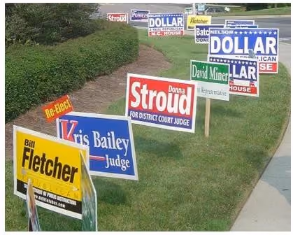 Campaign signs for local candidates on a grassy roadside