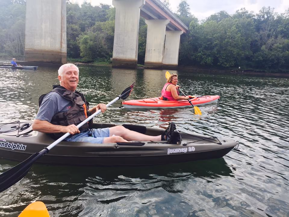 A man paddling a kayak on a river below a highway bridge, another kayaker behind him