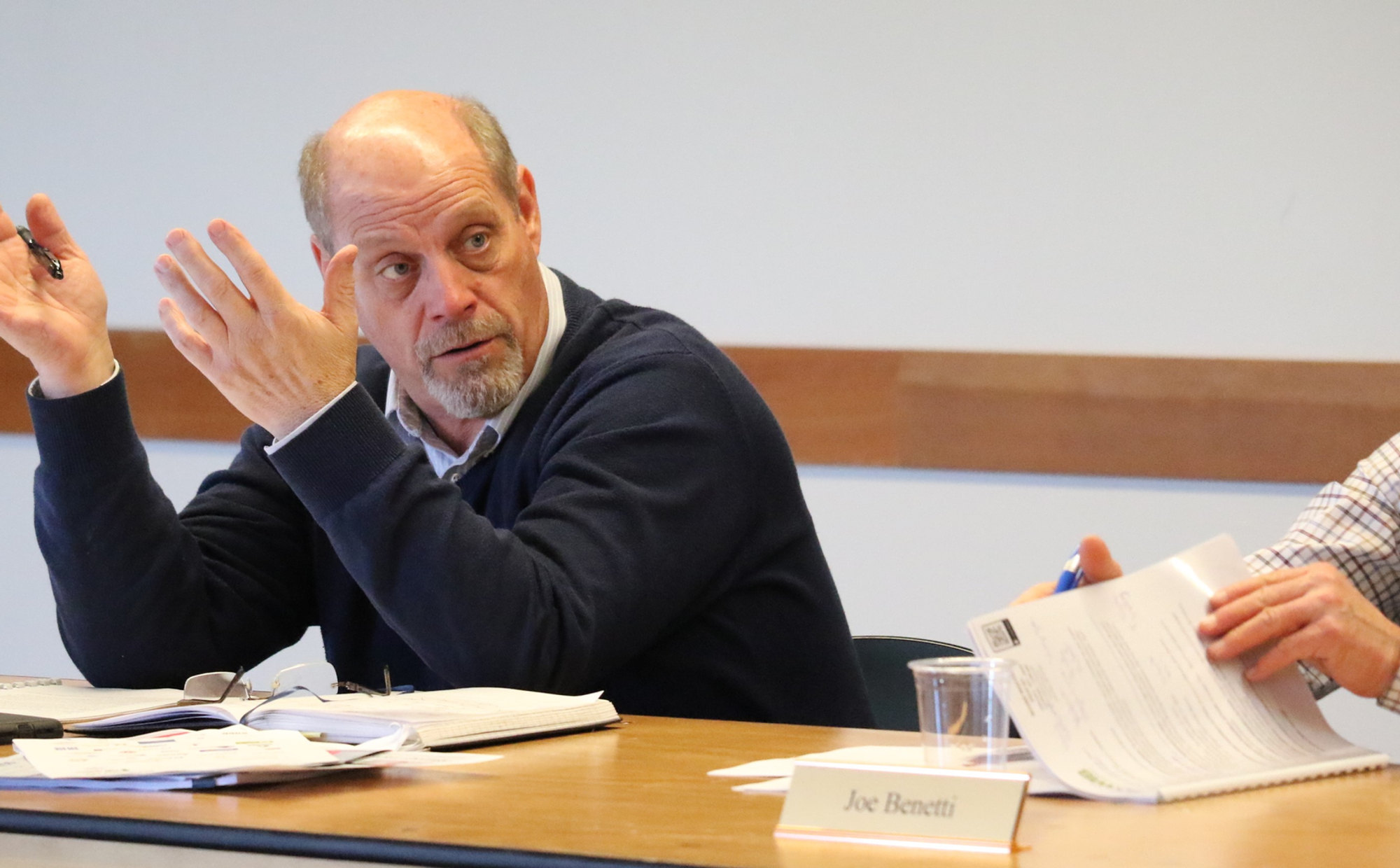 A board member speaks and gestures during a SOWIB board meeting, with agenda papers and a nameplate on the table in front of him.