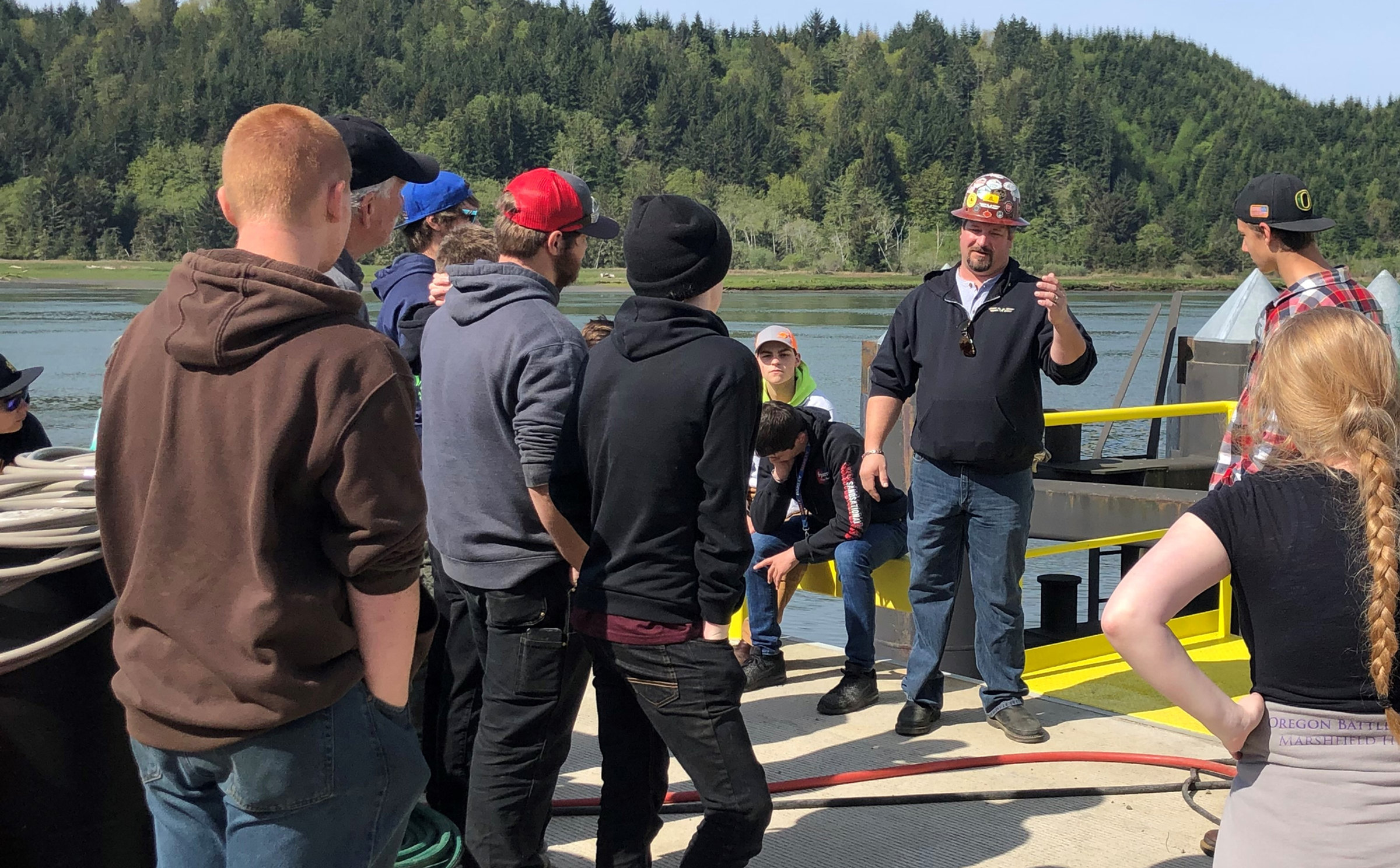 An instructor in a hard hat briefs a group of young people on a work barge on the estuary, with forested hills behind.