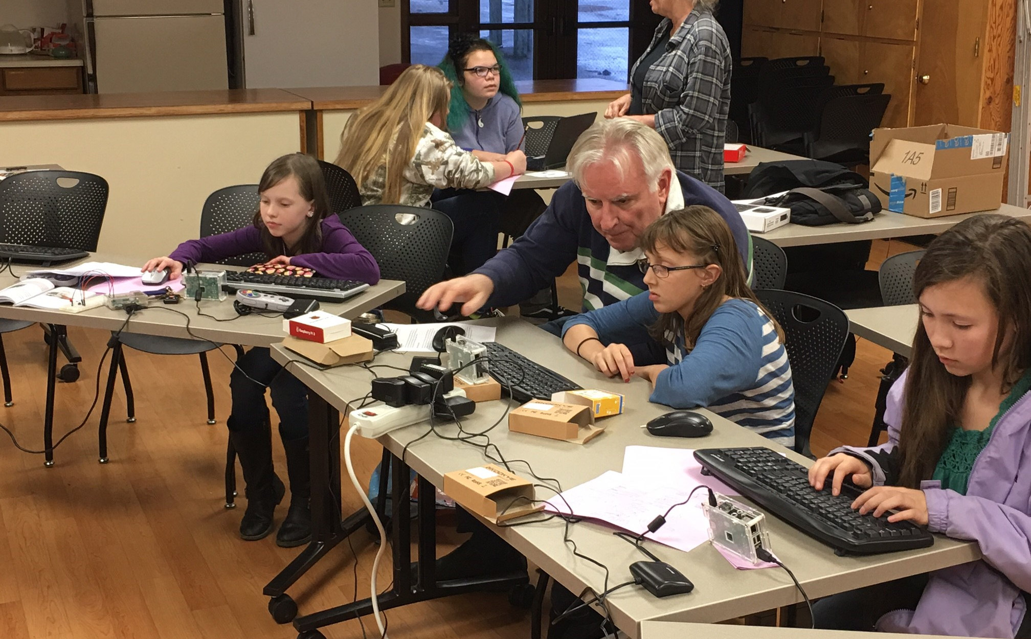 An instructor leans in to help a student at a hands-on electronics class, with single-board computers, keyboards and wiring laid out along the tables.