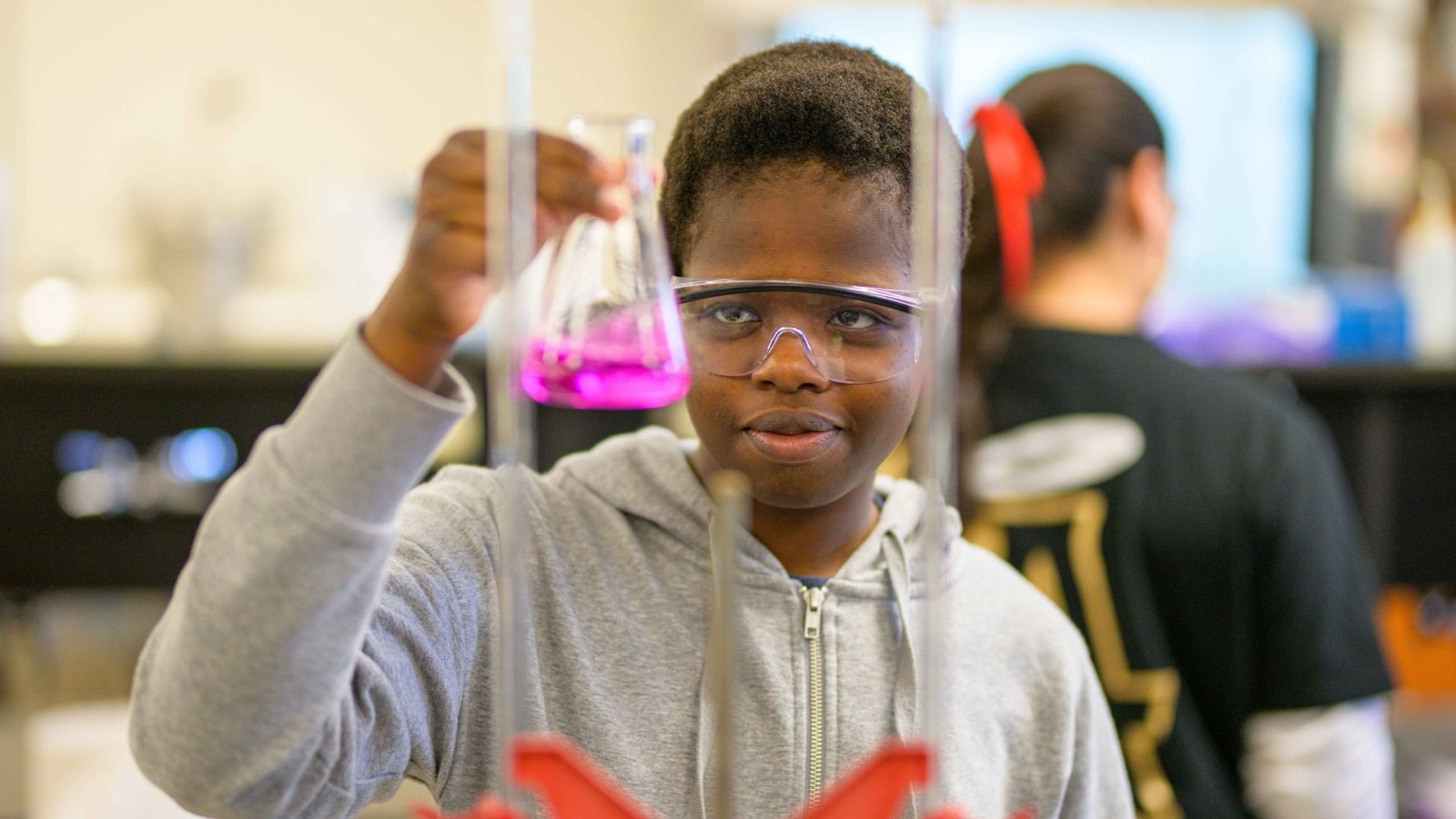 A young student in safety goggles raising an Erlenmeyer flask of bright pink liquid in a chemistry lab.