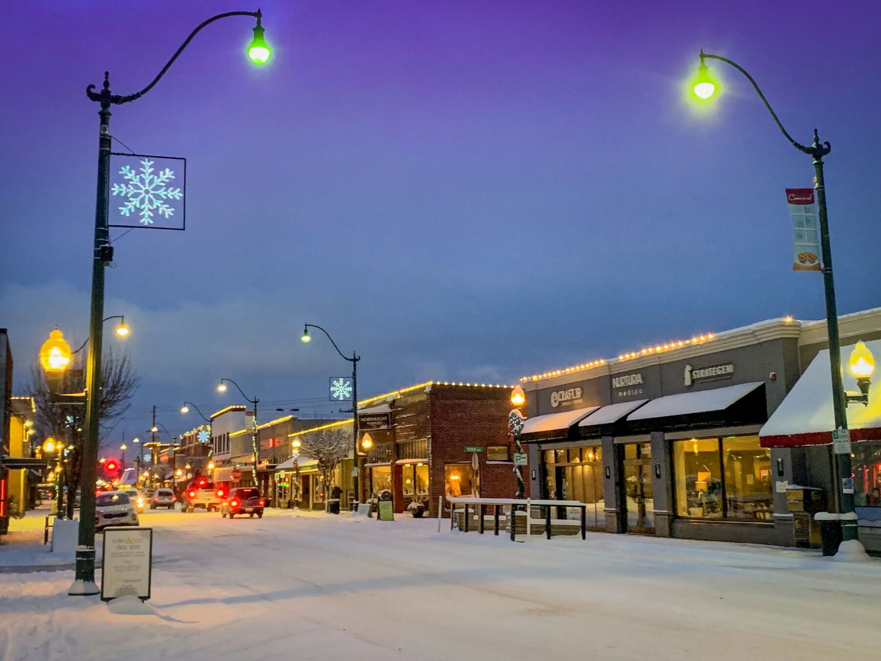 Main Street storefronts lit up at blue-hour dusk with fresh snow on the sidewalks and street.