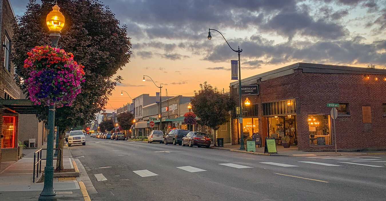 Main Street at summer sunset, hanging flower baskets glowing under lamplight outside brick storefronts.