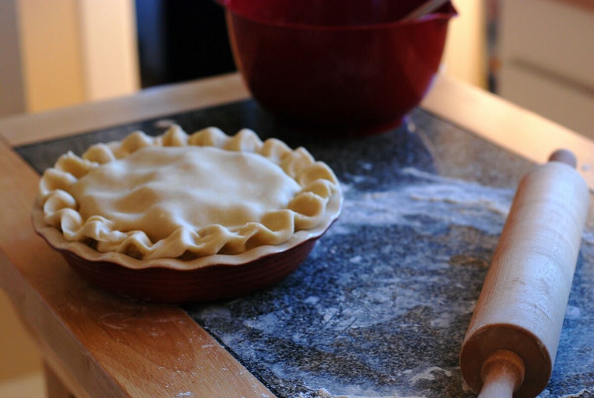 A hand-crimped pie crust ready for the oven on a flour-dusted counter beside a rolling pin.