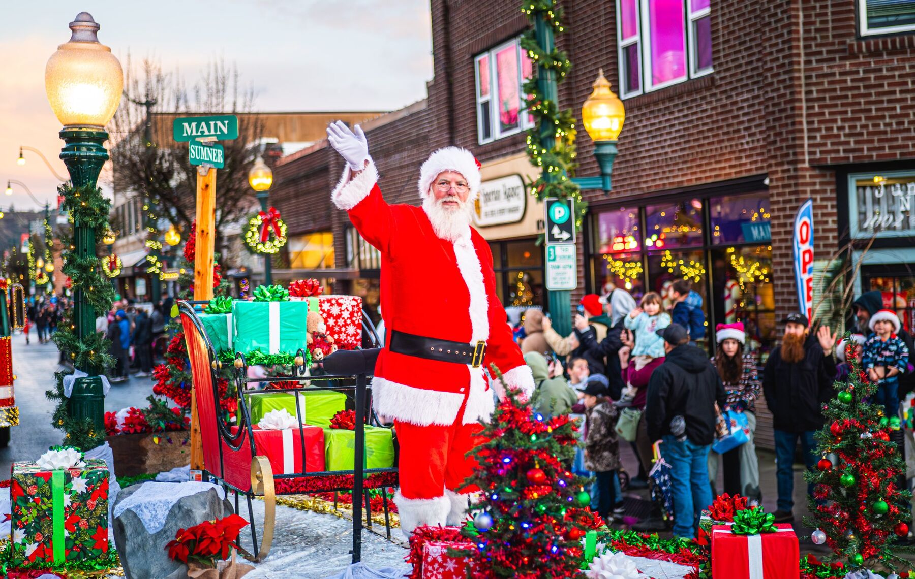 Santa waves from a sleigh float during the holiday parade down Main Street, the MAIN/SUMNER street sign visible.
