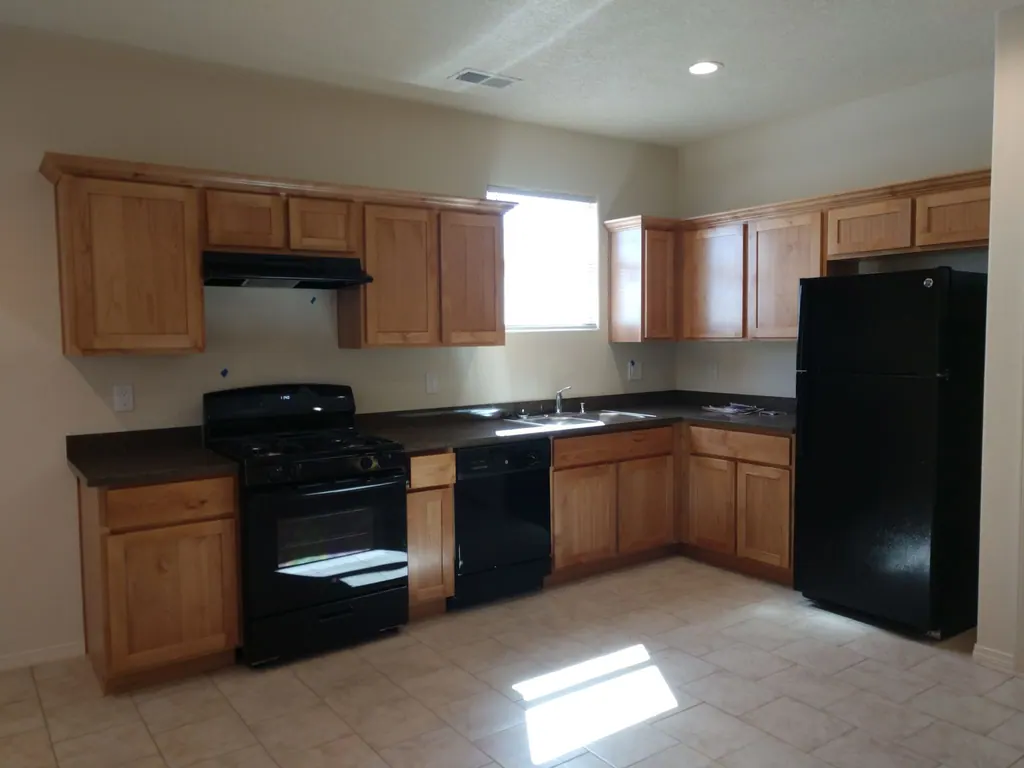 The kitchen of a new THI home with wood cabinets, a black range, refrigerator and dishwasher, and sunlight across a tile floor.
