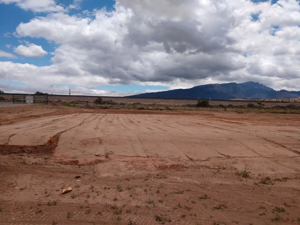 A graded red-earth lot before construction, with the Sandia Mountains on the horizon under a cloudy sky.