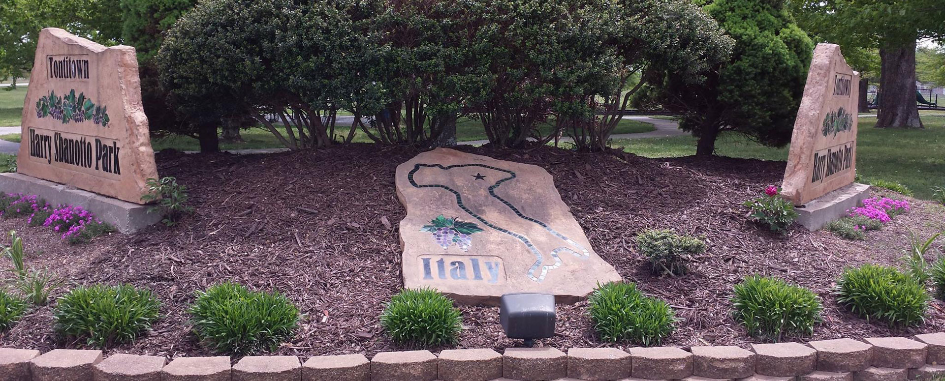 A stone marker at Harry Sbanotto Park with a mosaic map of Italy and grape clusters, flanked by the park's grape-carved entrance signs.