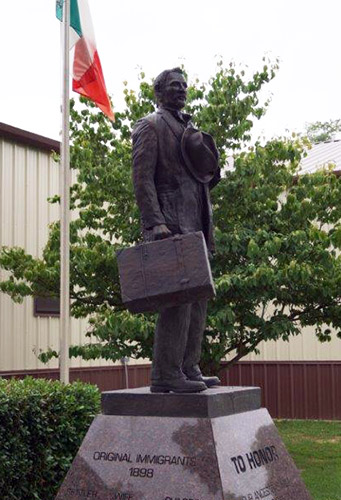 Bronze statue of an Italian immigrant in a suit holding a suitcase and hat, with the Italian flag flying behind it, honoring Tontitown's original 1898 settlers.