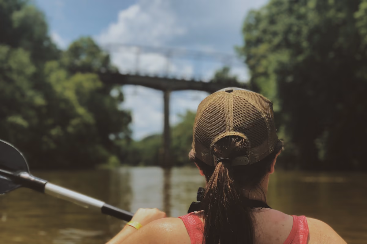 A woman in a ball cap paddling a kayak on the Duck River toward an old iron bridge.