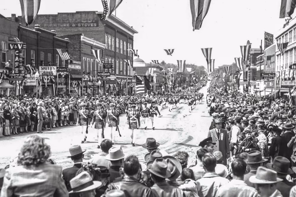 A black-and-white photograph of a marching band leading an early Mule Day parade through packed downtown streets hung with bunting.