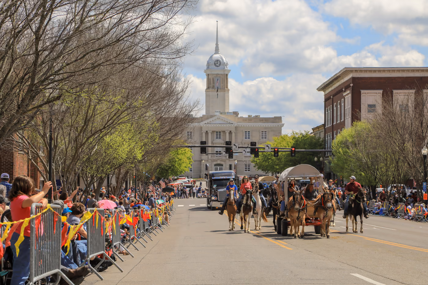 Riders and a covered wagon coming down the street toward the Courthouse during the Mule Day parade, with crowds along the barricades.