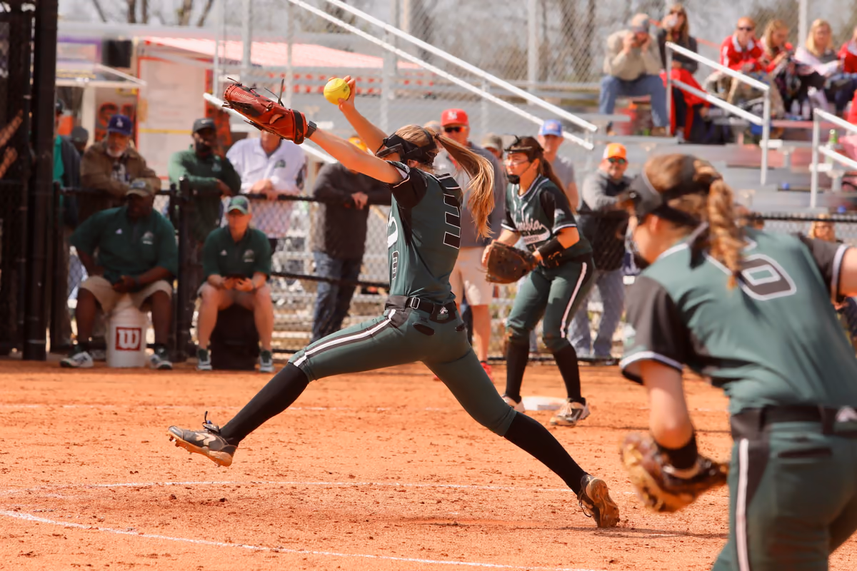 A softball pitcher in green mid-windmill delivery during the Midstate Classic, with teammates and fans behind her.
