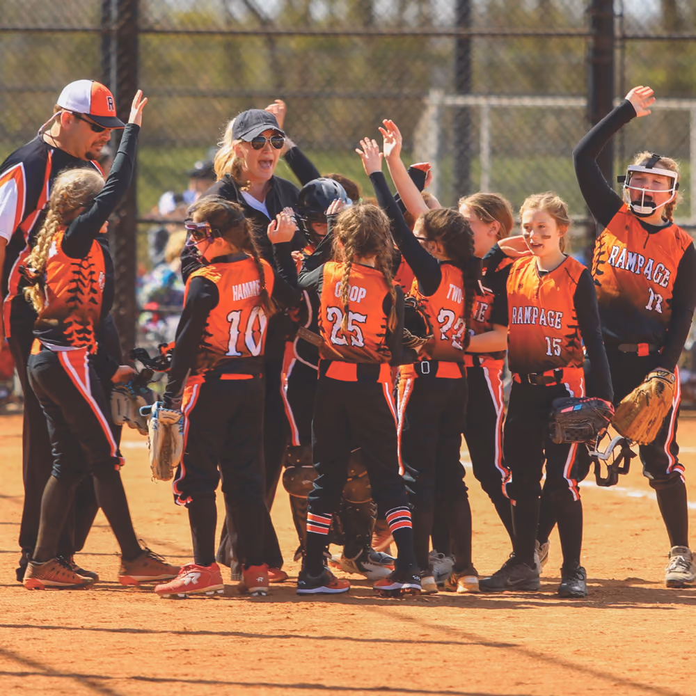 A girls' softball team in orange and black jerseys celebrating together on the infield.