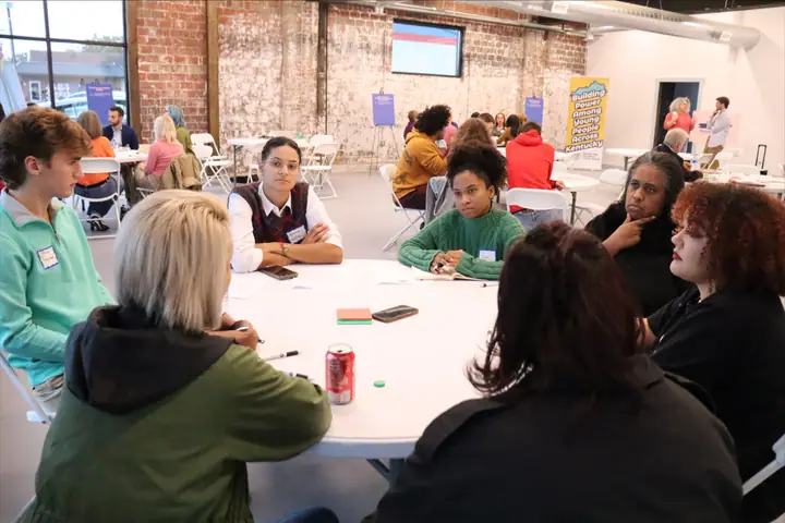 Students and adults talk around a round table in a brick-walled room.