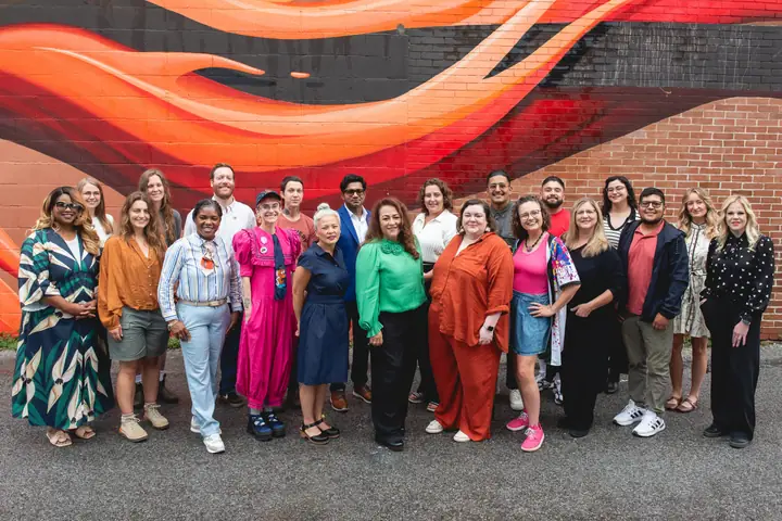 About two dozen people stand together in front of a painted orange and red mural on a brick wall.