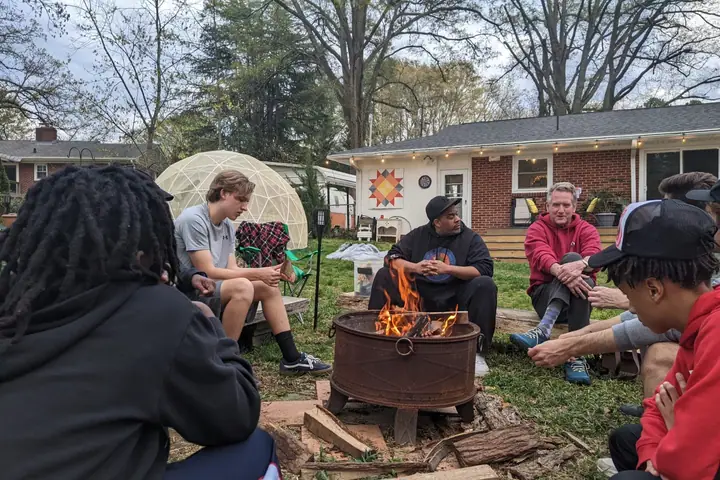 Teenage boys and adult mentors sit in a circle around a fire pit in a back yard.