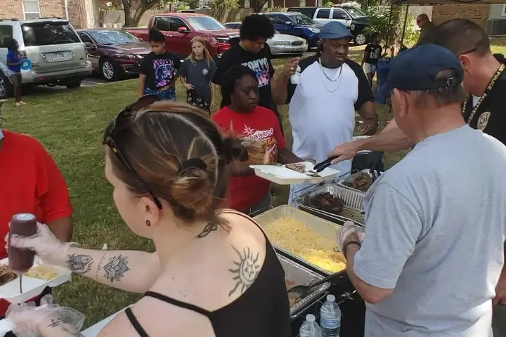 Volunteers in gloves serve barbecue to neighbors from a table outdoors.