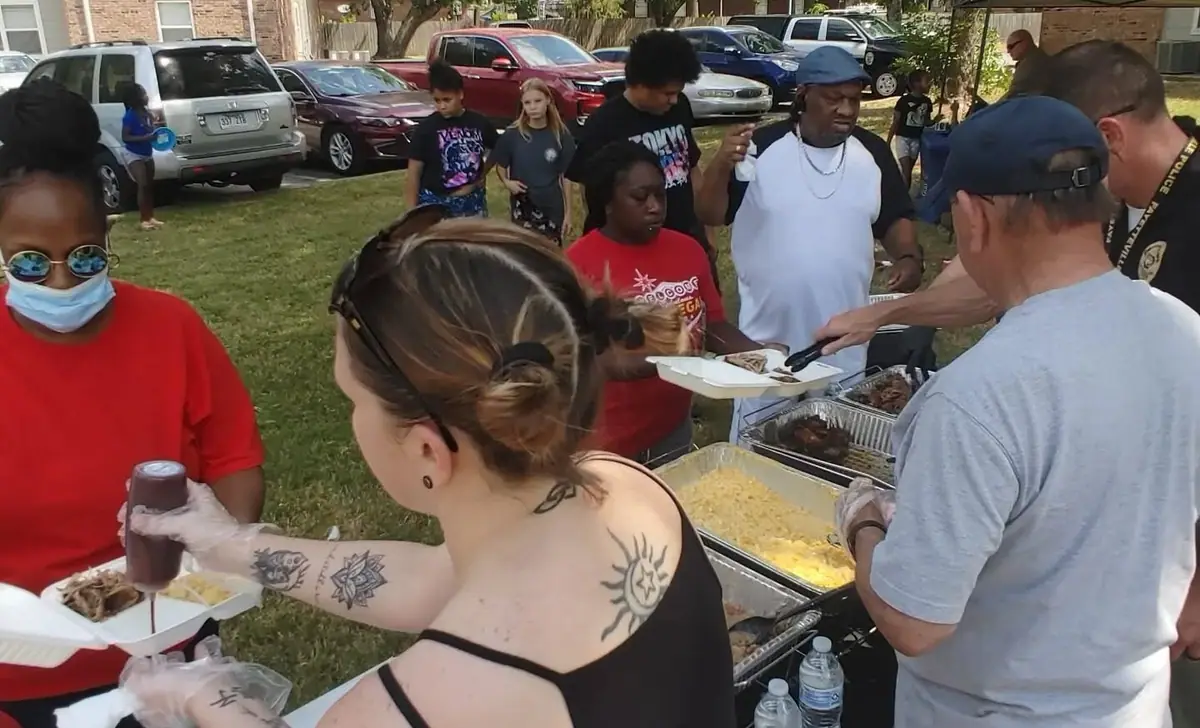 Volunteers in gloves serve barbecue to neighbors from a table outdoors.