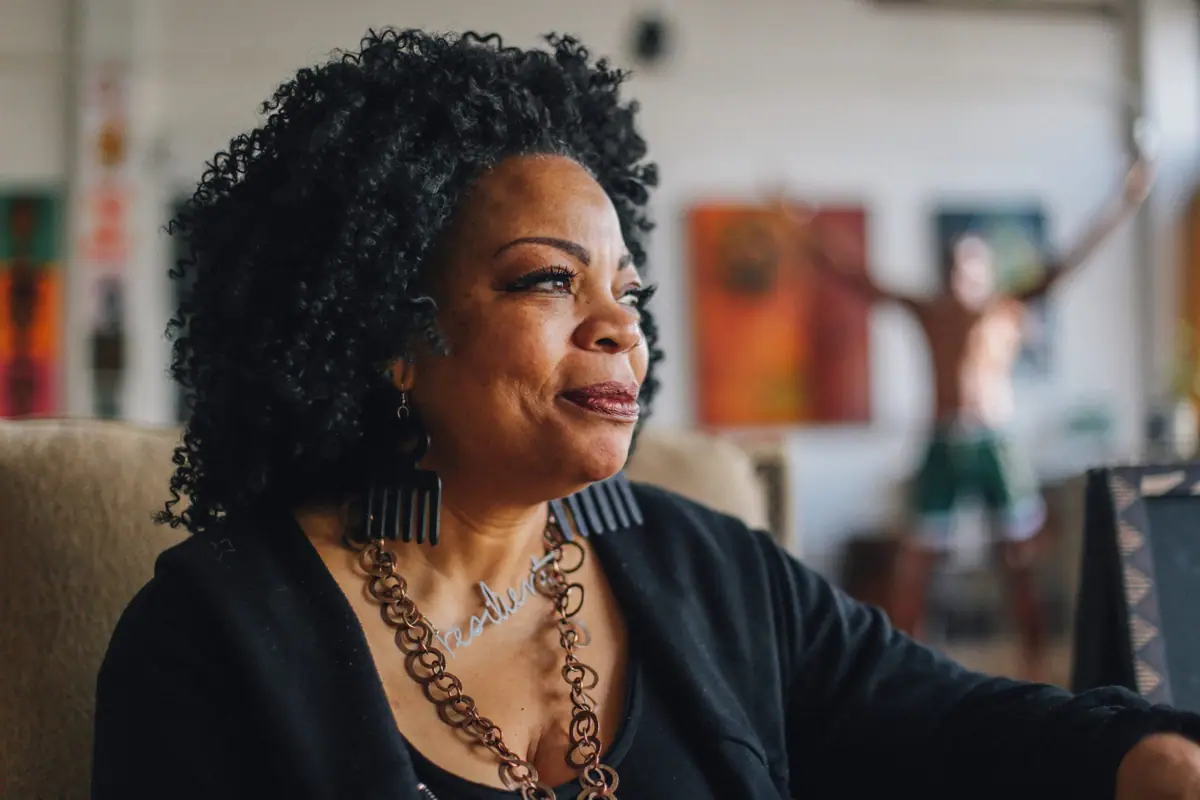 A woman with curly hair and a beaded necklace looks across a room hung with artwork.