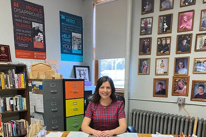 A teacher sits at her desk under a wall of framed portraits; a pennant banner on the desk reads Mrs. Helberg.