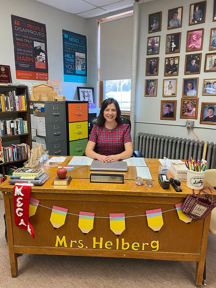 A teacher sits at her desk under a wall of framed portraits; a pennant banner on the desk reads Mrs. Helberg.