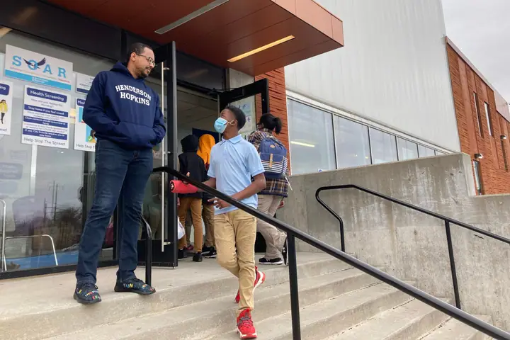 A man stands at the entrance of a school as a masked student walks down the steps past him.