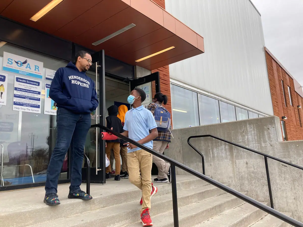 A man stands at the entrance of a school as a masked student walks down the steps past him.