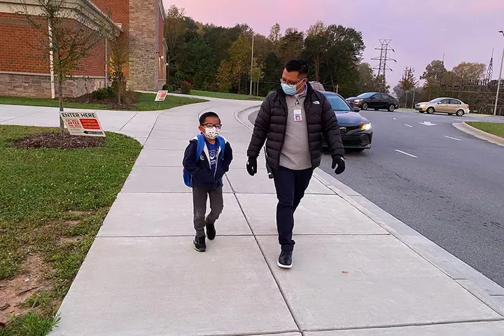 A man and a boy in face masks walk along a sidewalk outside a school at dusk.