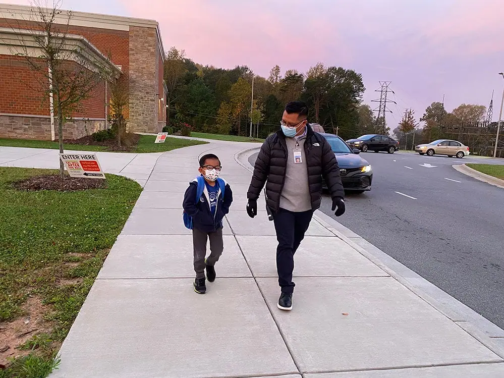 A man and a boy in face masks walk along a sidewalk outside a school at dusk.